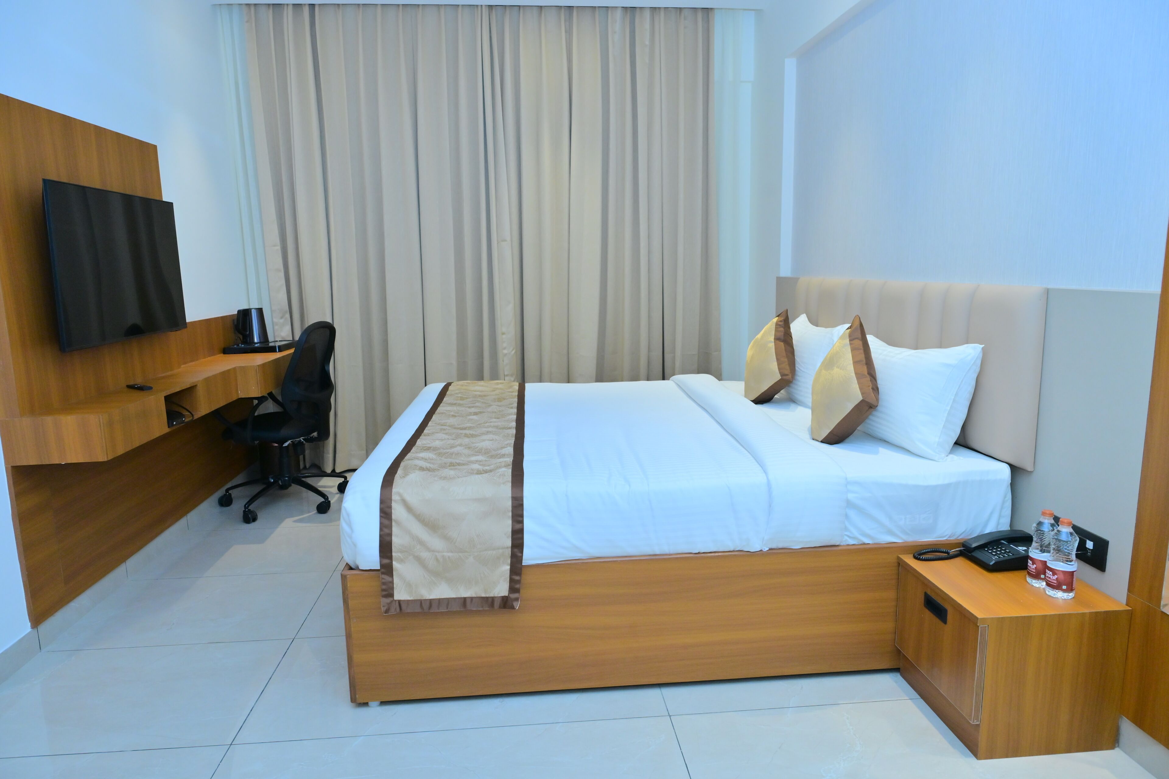 A hotel room with a bed, a desk with a chair, a television, and a nightstand with a phone and water bottles.