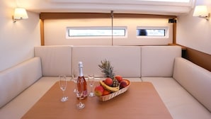 Schiffsinnenraum mit gepolsterten Sitzgelegenheiten, einem Tisch mit Flasche und Gläsern sowie einem Obstkorb.