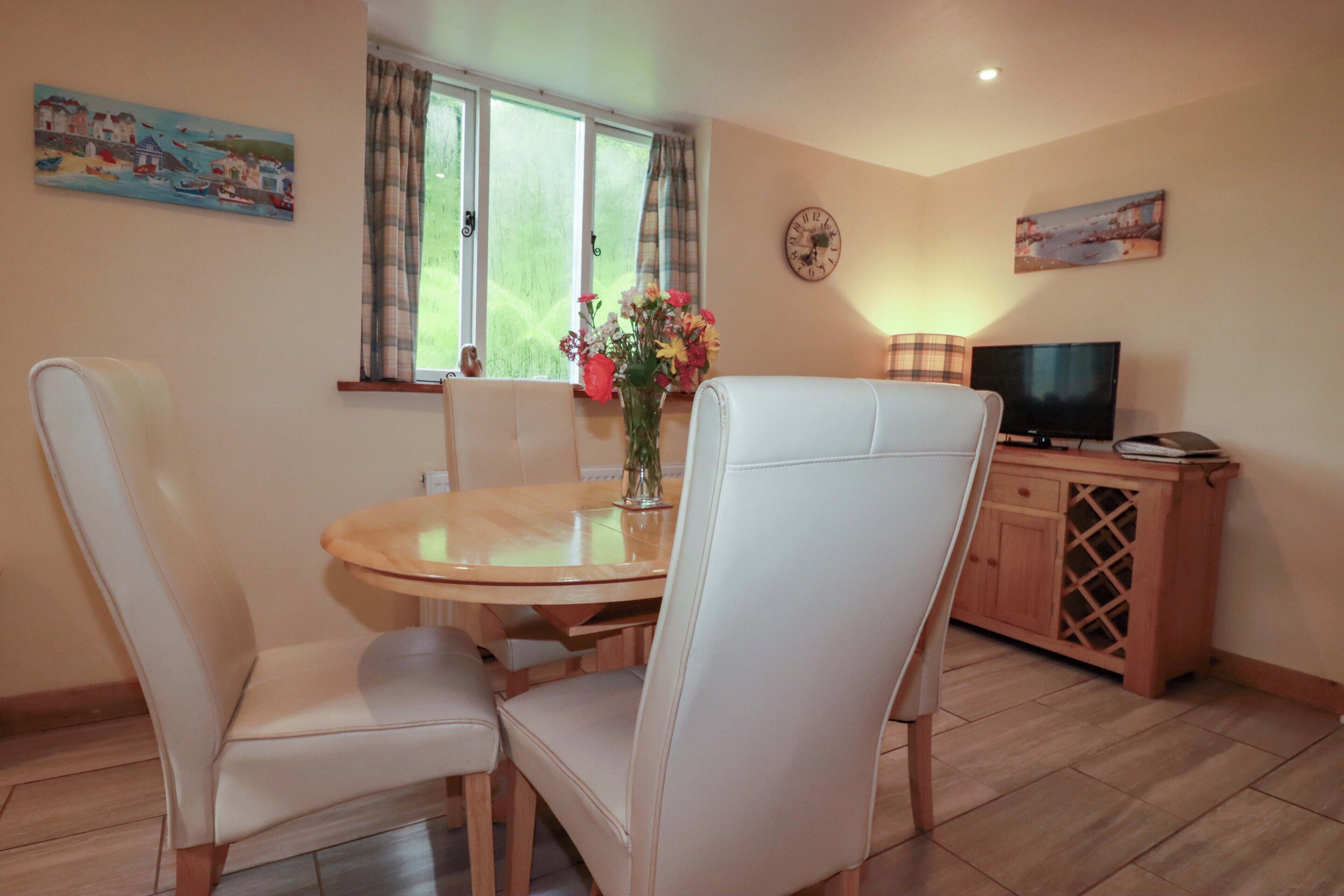 A dining area with a round table, four white chairs, a vase with flowers, a clock on the wall, and a television on a wooden cabinet.