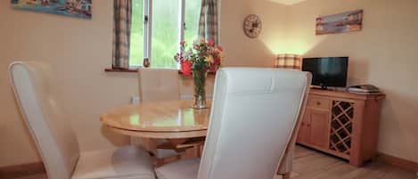 A dining area with a round table, four white chairs, a vase with flowers, a clock on the wall, and a television on a wooden cabinet.