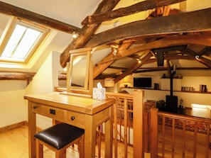 Un escritorio de madera con un espejo, una silla negra, una mesita pequeña y un televisor en una habitación estilo loft con vigas a la vista y un tragaluz.