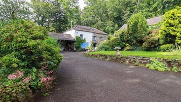 A gravel driveway leading to a stone house with a blue door, surrounded by lush greenery and a stone wall.