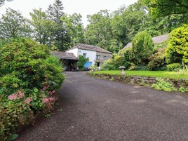 A gravel driveway leading to a stone house with a blue door, surrounded by lush greenery and a stone wall.