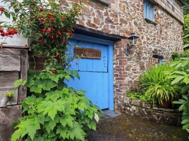 Un edificio rústico de piedra con una puerta azul, un letrero de madera encima de la puerta y una planta en maceta a la derecha.