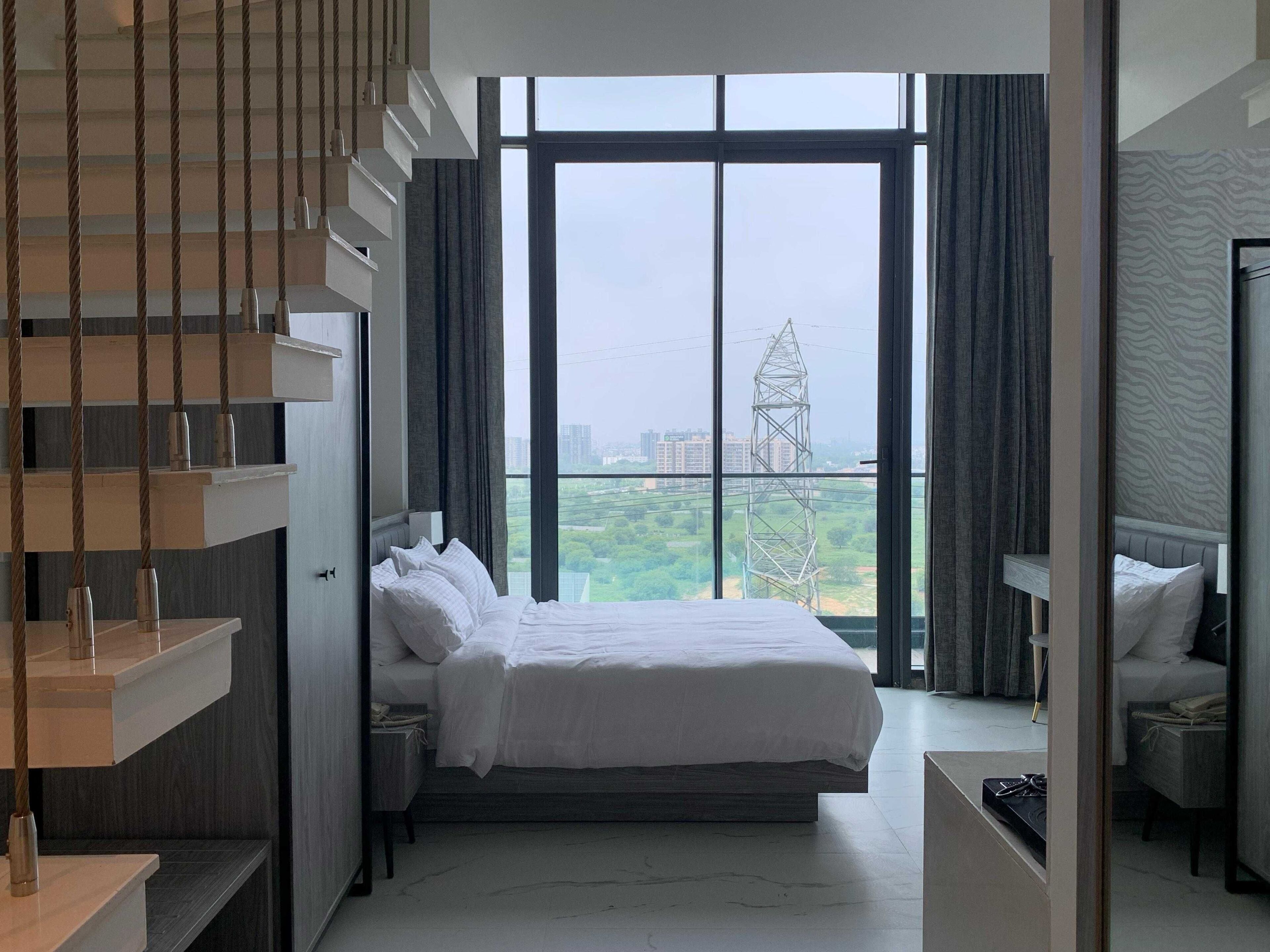 A modern hotel room with a large bed, a built-in desk, a staircase, and a view of the cityscape.