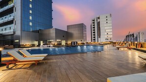 Un área de piscina en la azotea con sillones de descanso, una piscina y edificios modernos al fondo.