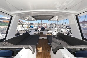 Convés de barco com assentos, um leme e vista para um porto com outros barcos.