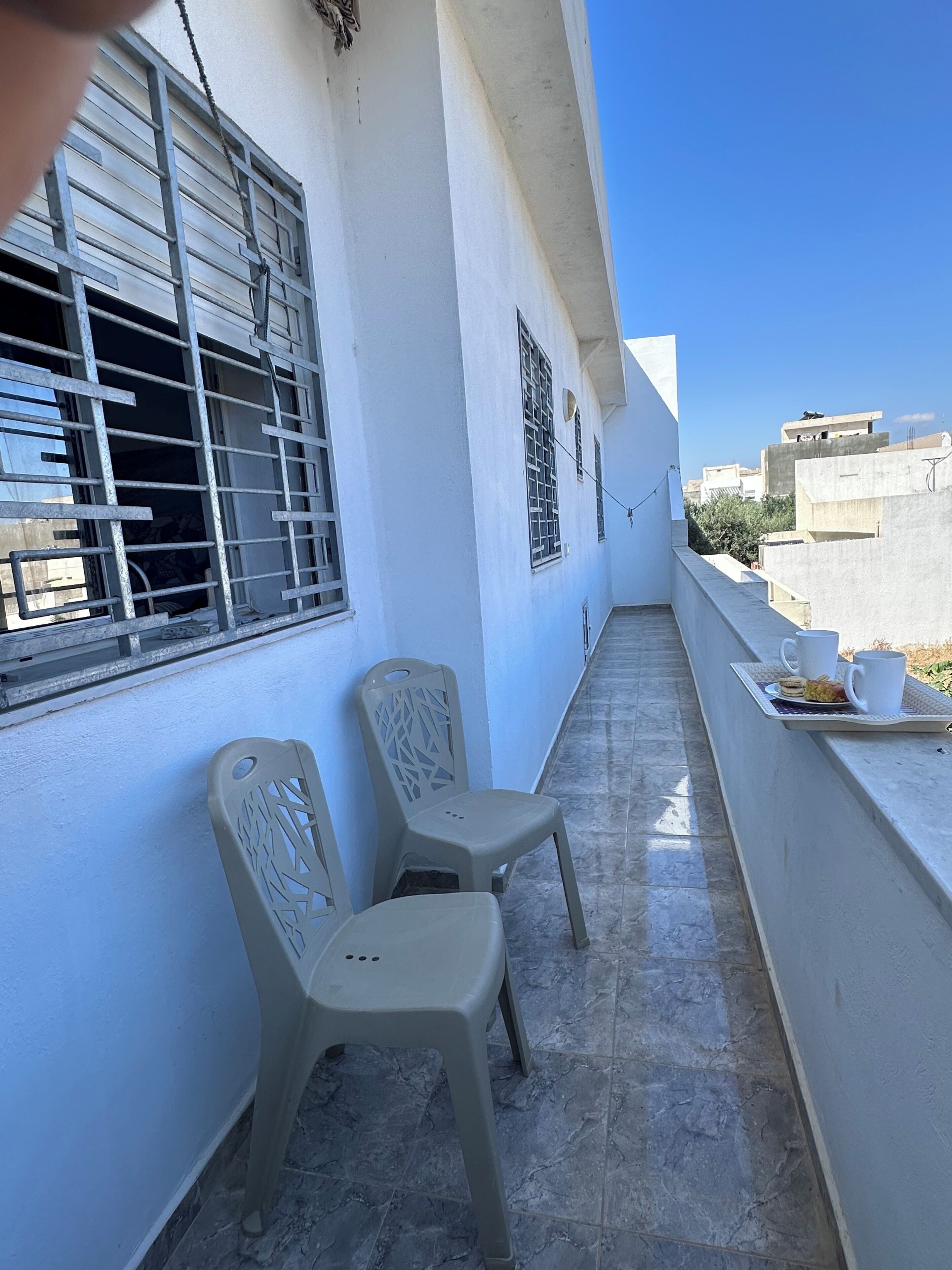 A balcony with two plastic chairs, a tray with cups and a pastry, and a view of buildings in the background.