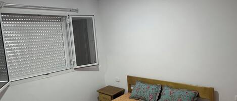 Une chambre à coucher comprenant un lit, deux tables de chevet, une fenêtre avec des stores et un luminaire au plafond.