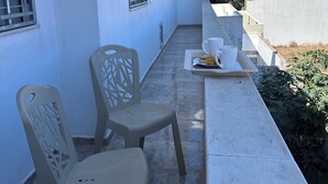 Un balcón con dos sillas de plástico, una mesa con una taza y plato, y un ventanal con barrotes.