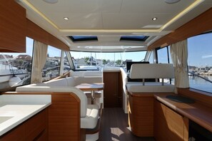 Interior de barco com assentos, uma mesa e vista para o porto de iates.