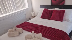 A neatly made bed with white and red bedding, a red rose painting on the wall, and a window with blinds. - Elysea Haven 3 Bedroom house, newly renovated (Leeds)