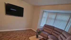 A living room with a large sectional sofa, a flat-screen TV mounted on the wall, a wooden herringbone floor, and a window with blinds. - Elysea Haven 3 Bedroom house, newly renovated (Leeds)