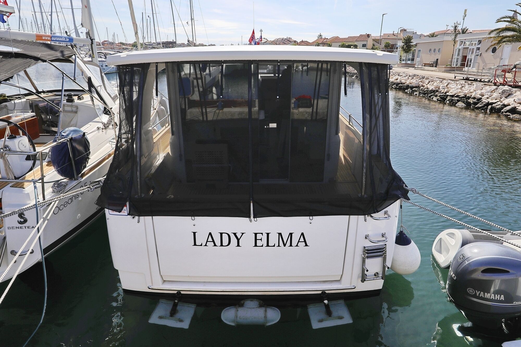 Ein Boot mit dem Namen 'Lady Elma' liegt in einer Marina festgemacht, mit weiteren Booten, einer felsigen Küste und Gebäuden im Hintergrund.
