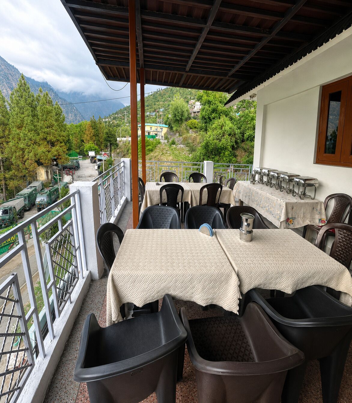 An outdoor terrace with tables and chairs, a railing, and a view of mountains and parked vehicles.