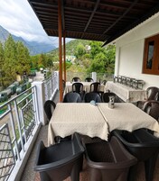 Una terraza al aire libre con mesas y sillas, barandilla y vistas a montañas y vehículos estacionados.