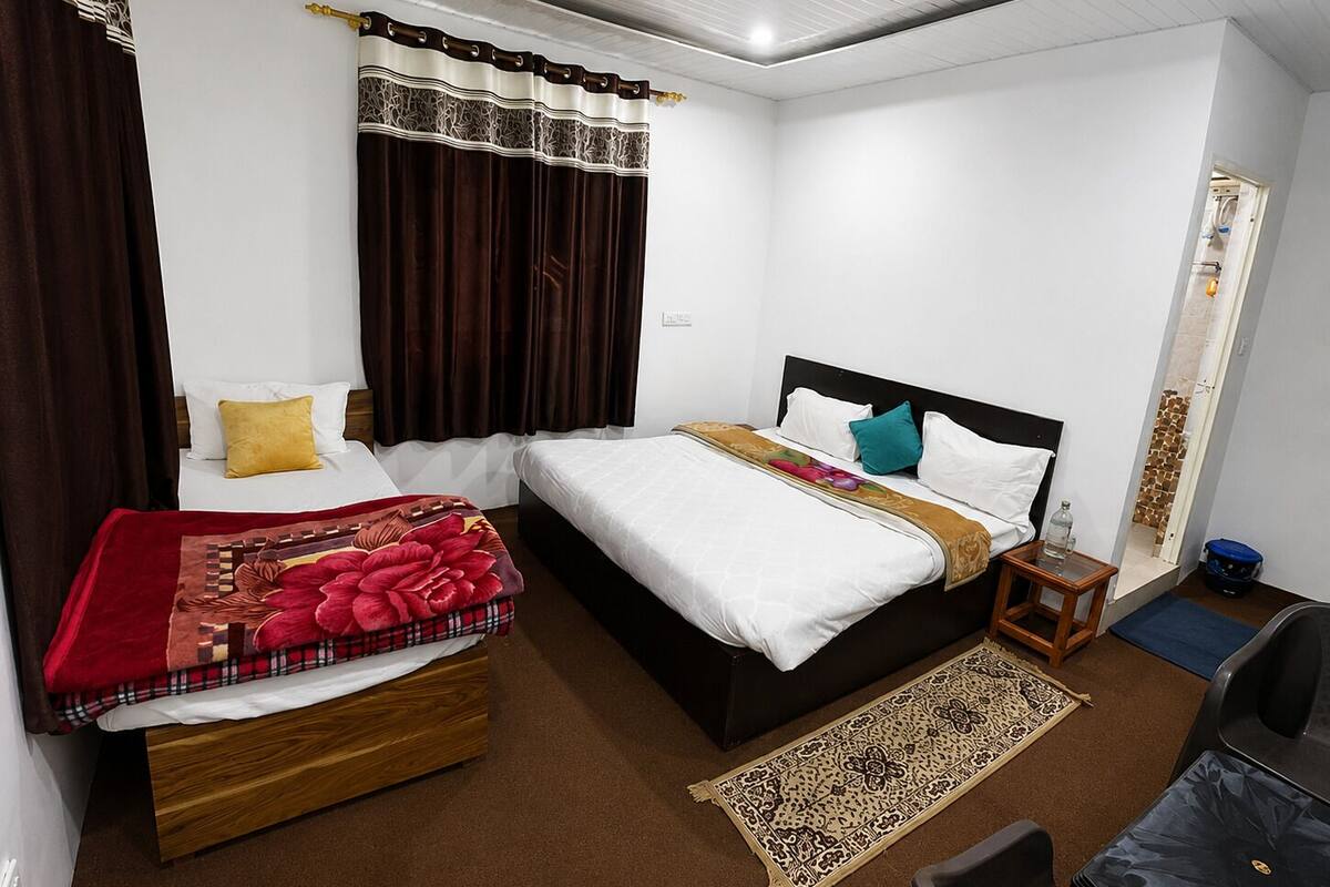 Une chambre d’hôtel avec deux lits, une tête de lit en bois, une table de chevet, une petite table, une chaise, un espace douche et un tapis à motifs.