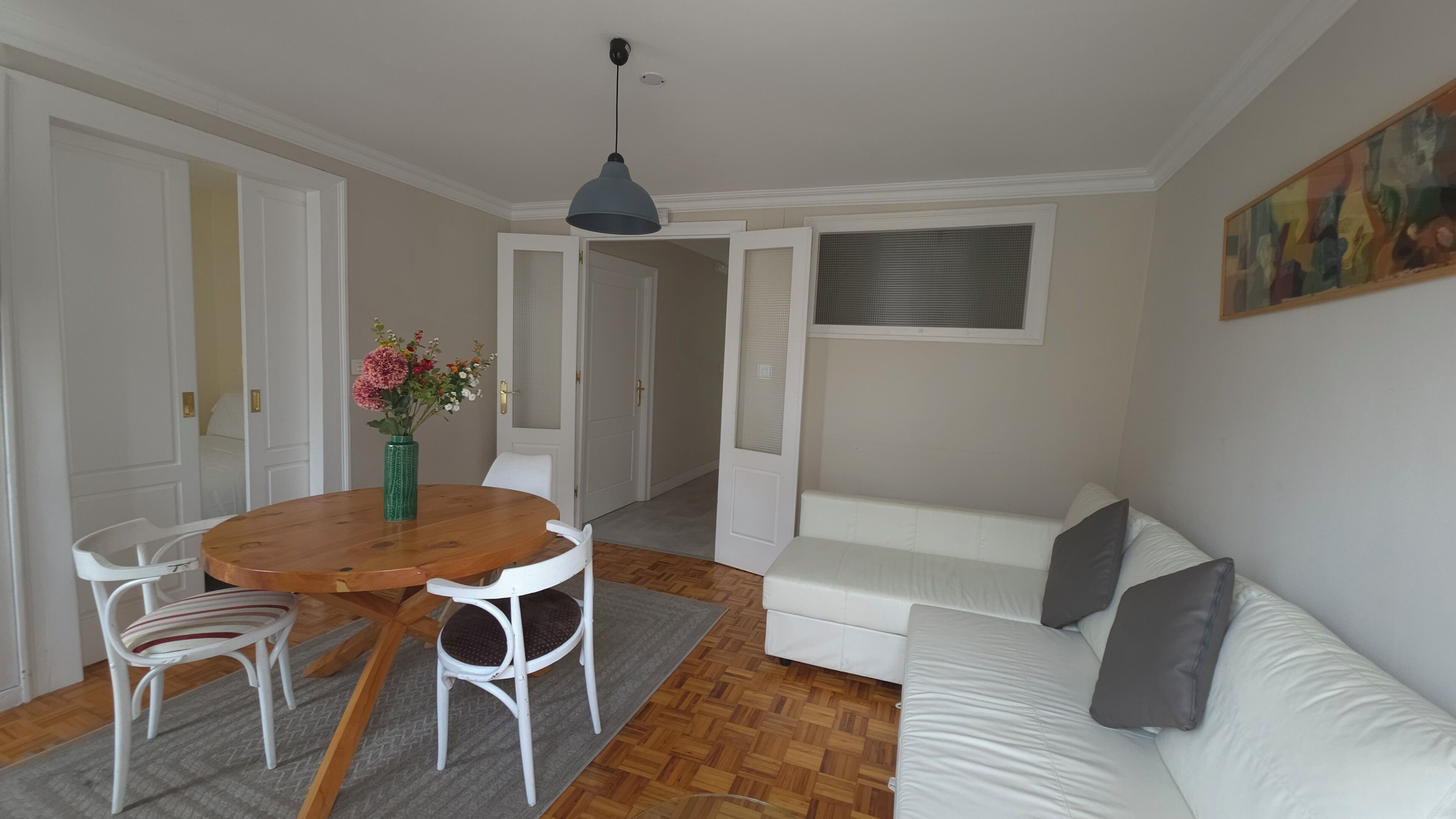 Een kamer met een ronde houten tafel, twee witte stoelen, een witte bank, een grijze vloerkleed en een hangende lamp, plus een schilderij aan de muur.