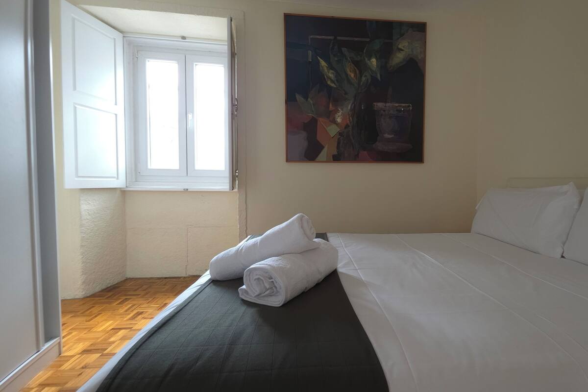 A hotel room with a bed, a window, and a painting on the wall.