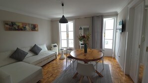 Una sala de estar con un sofá blanco, una mesa de comedor redonda de madera, una silla blanca, una mesita blanca y un lámpara colgante.
