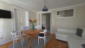Una habitación con una mesa de comedor de madera, sillas blancas, un sofá blanco, un tapete gris y una lámpara en el techo.