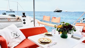 Interior de barco con mesa de comedor, cojines naranja, almohada blanca con un logo, vaso de agua, plato de comida y florero con flores.