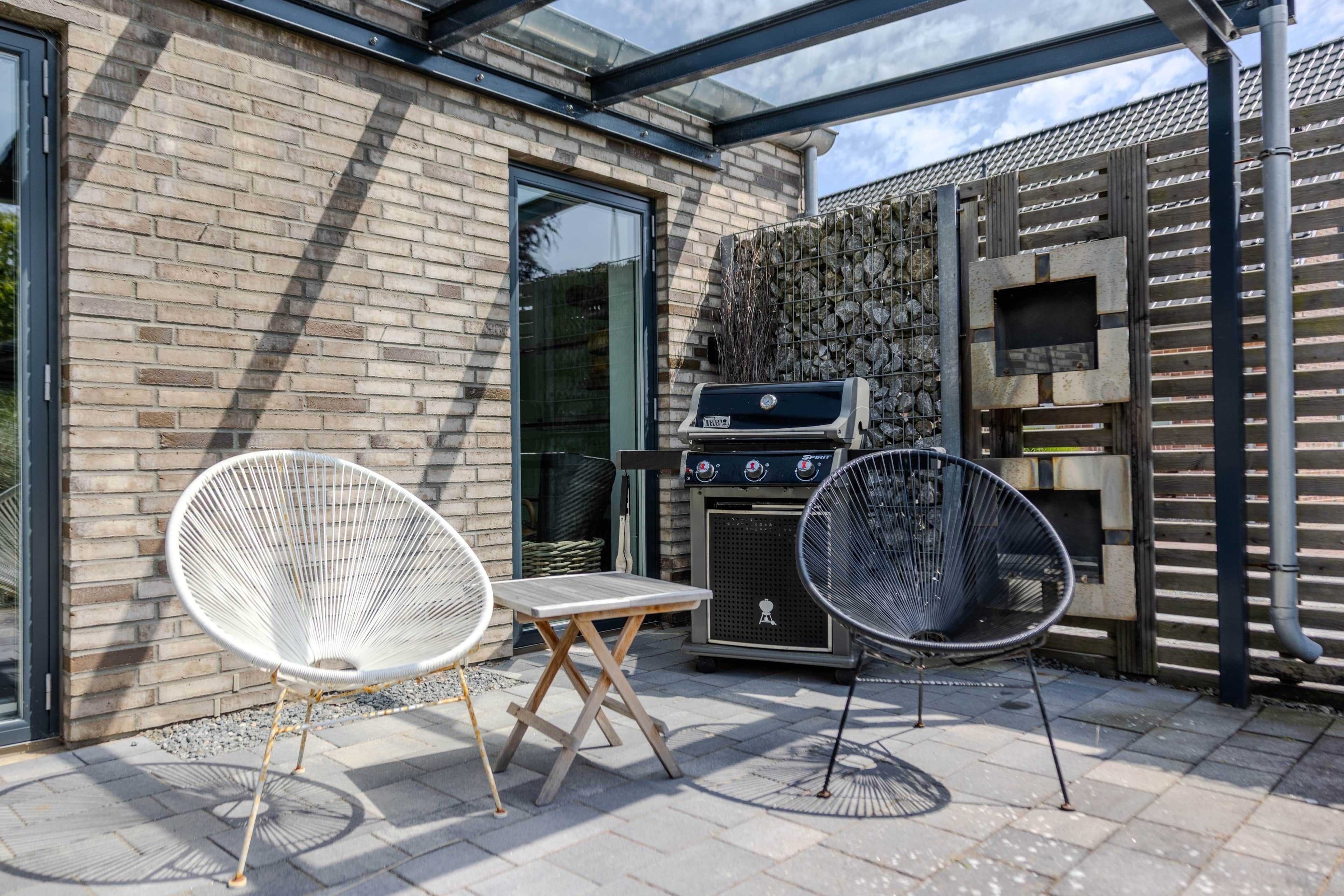 An outdoor patio with two white wire-framed chairs, a small wooden table, a barbecue grill, and a pizza oven.