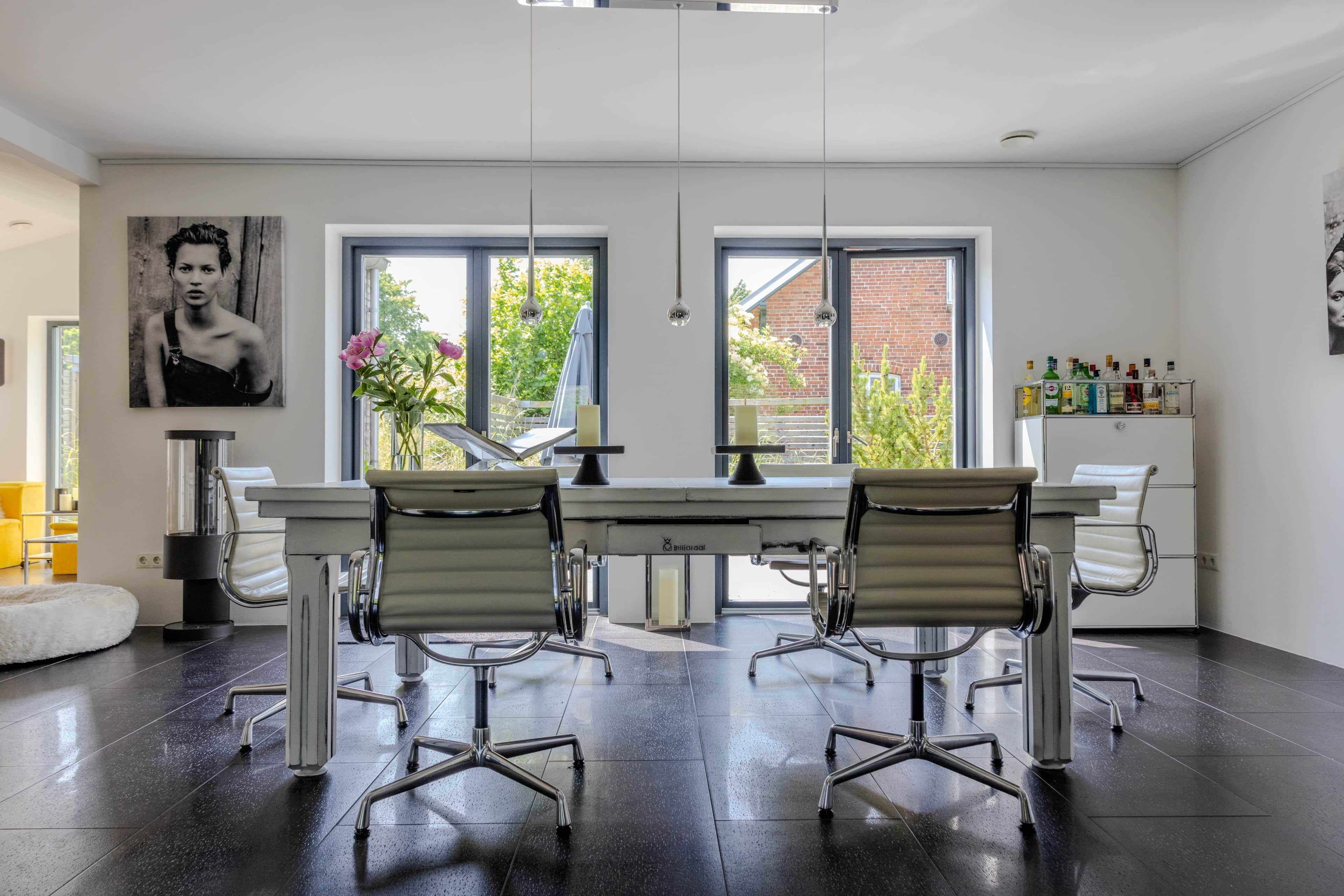 Un espace de bureau moderne avec une grande table, deux chaises de bureau, un canapé blanc et une grande fenêtre donnant sur un paysage verdoyant.