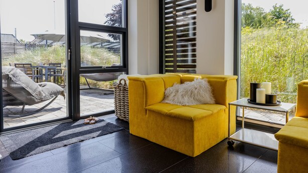 Una sala de estar moderna con un sofá amarillo, una mesa de centro de vidrio y vistas a un jardín a través de ventanas amplias.