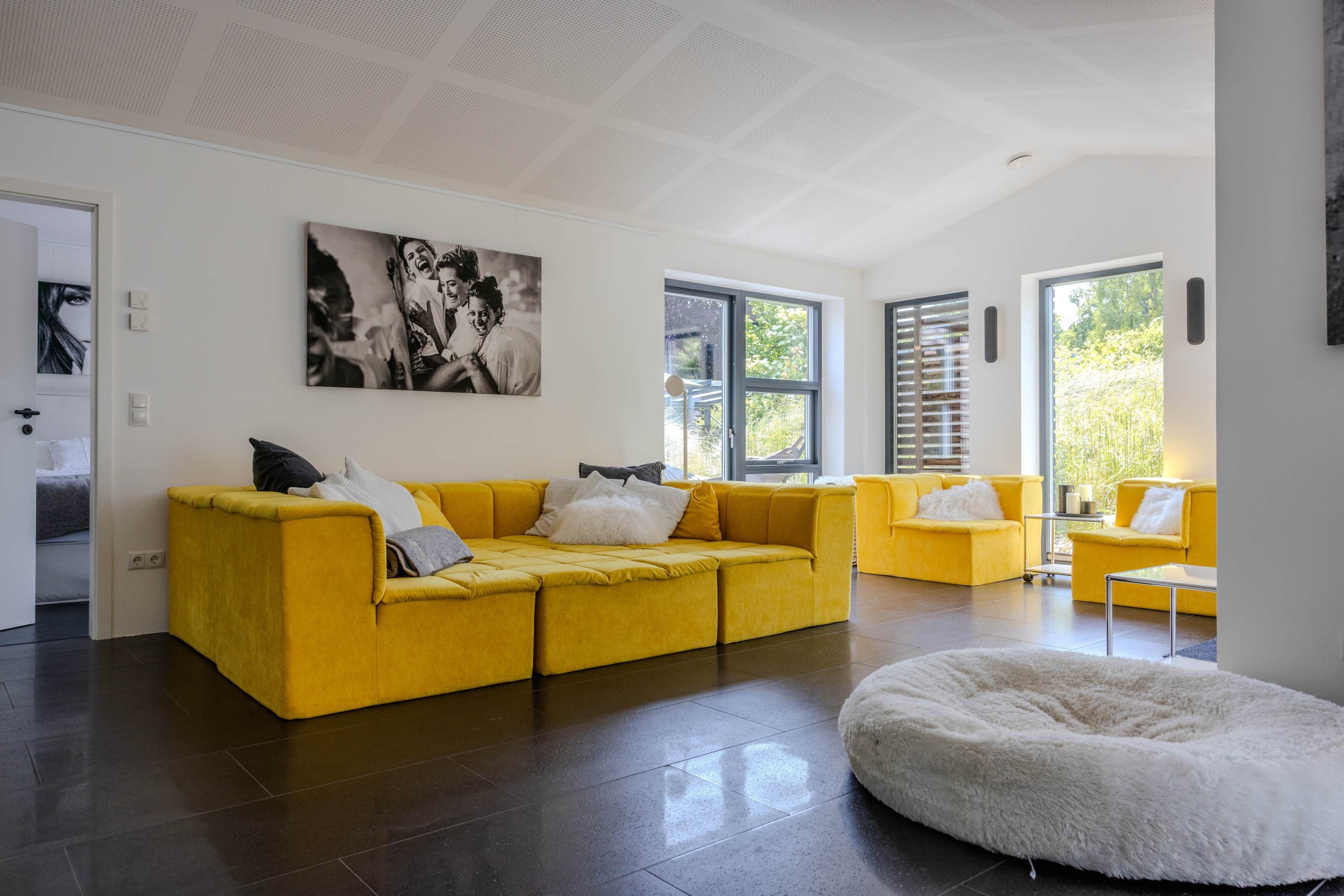 A modern living room with a yellow sofa, a white fluffy ottoman, and a large black-and-white painting on the wall.