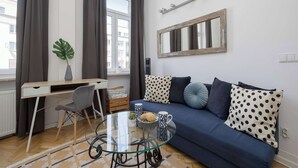 Uma sala de estar moderna com uma mesa de centro de vidro, um sofá azul com almofadas estampadas, uma escrivaninha com cadeira e uma janela grande com cortinas.