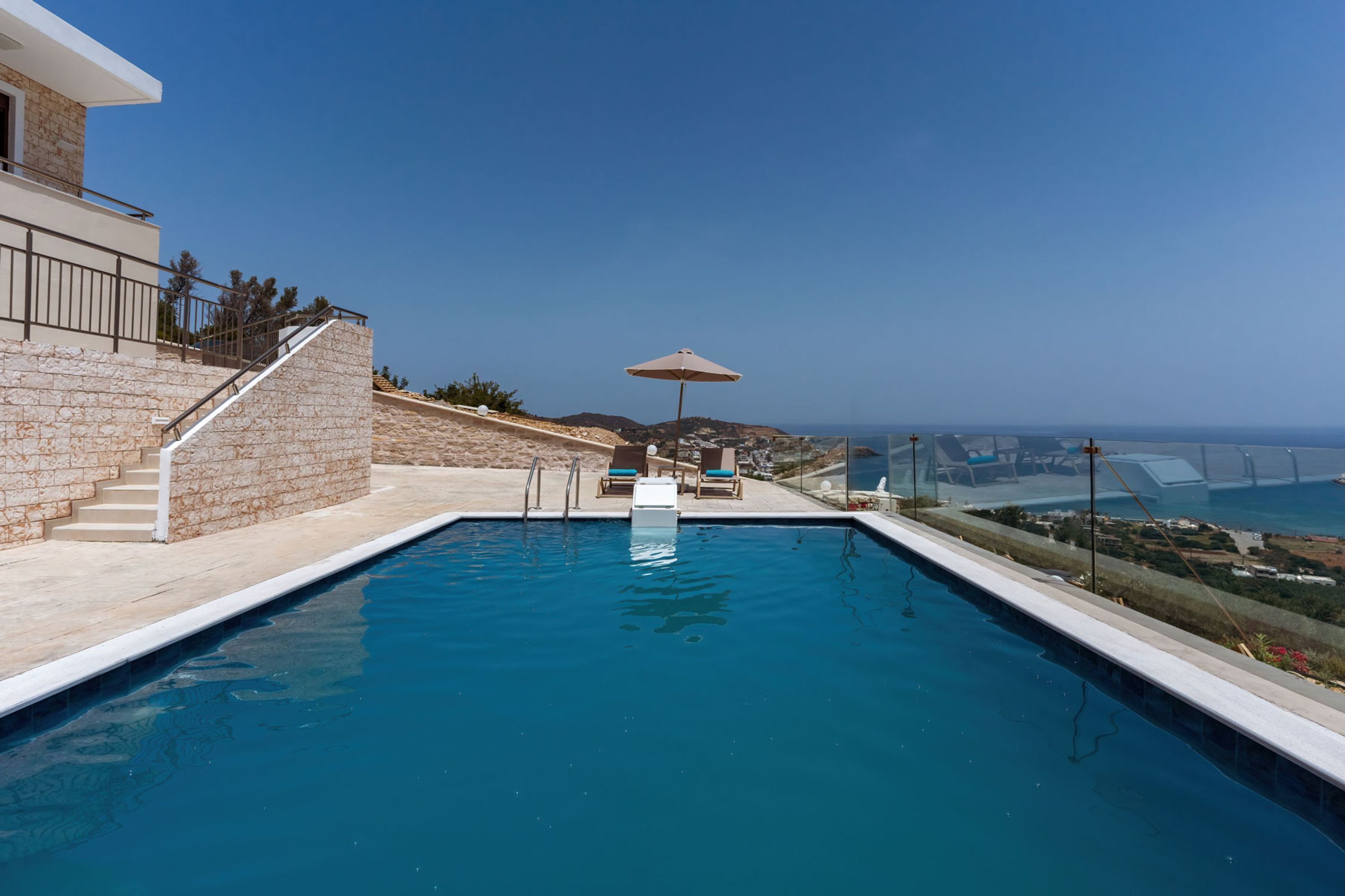 A swimming pool with a view of the sea, a staircase, and a lounge area with chairs and an umbrella.