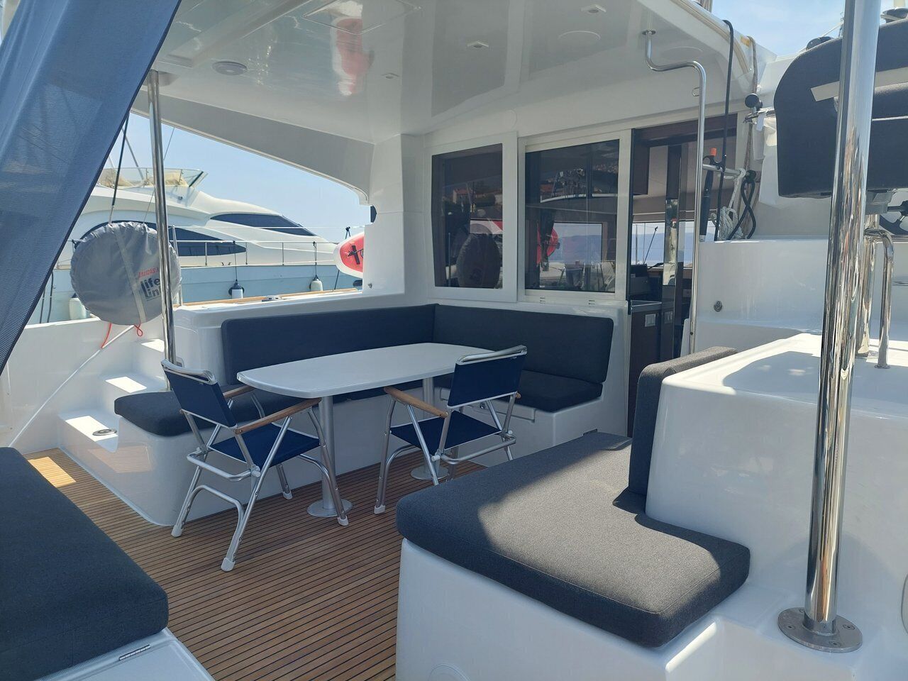 A boat's deck with a table and chairs, a cushioned seating area, and a window with a view of the sea.