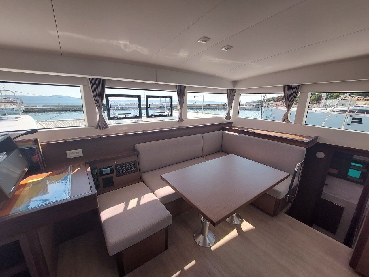 A boat interior with a control station, seating area, and windows offering views of the water and docked boats.