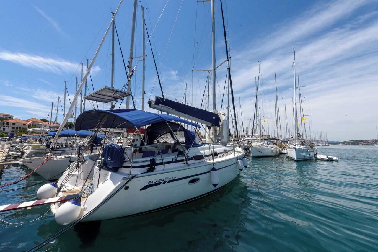Una marina con varios veleros amarrados, un área techada y edificios al fondo.
