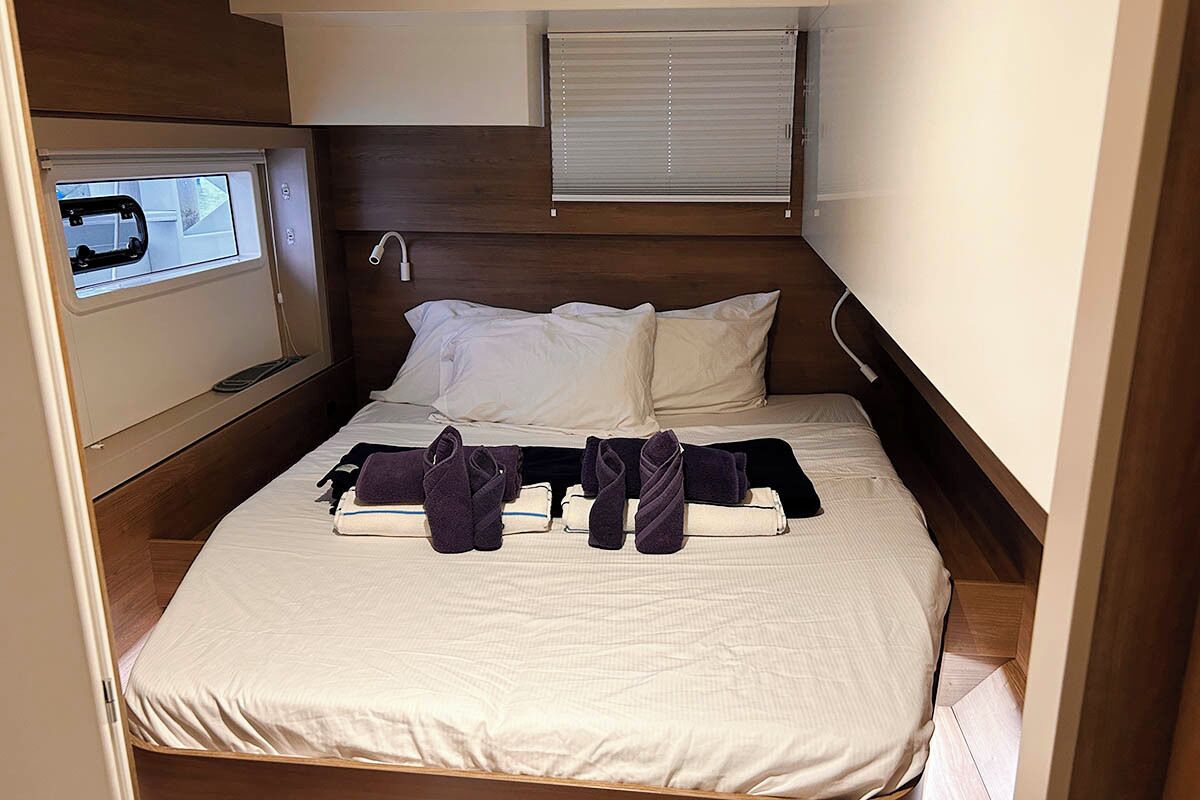 Un lit compact avec une tête de lit, une fenêtre avec des stores et une petite étagère avec des serviettes.