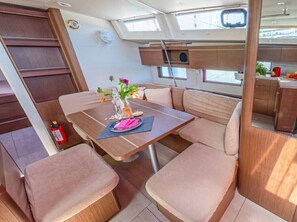 A boat interior with a dining area, cushioned seating, a small table with a vase of flowers, and a kitchen area with a sink and countertop.