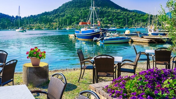Un ristorante sul lungomare con posti a sedere all'aperto, barche ormeggiate nelle vicinanze e una montagna sullo sfondo.