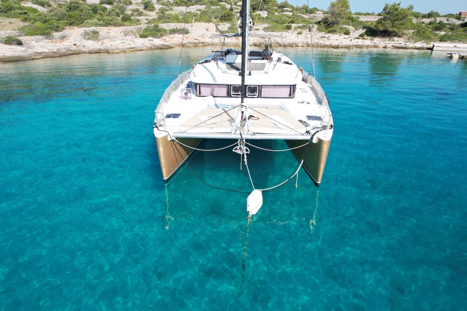 Ein weißes Katamaranboot liegt vor Anker in klarem türkisblauem Wasser, im Hintergrund felsiges Gelände.