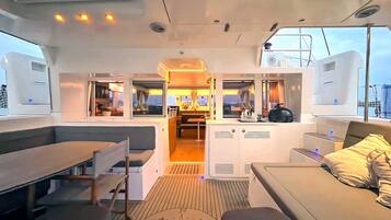 Interior de un barco con zona de comedor, cocina y área de estar con vista al mar.