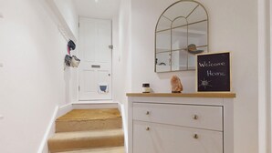 Un escalier recouvert d’une moquette beige mène à une porte blanche. On y trouve également un meuble blanc avec des tiroirs, un miroir et un tableau noir sur lequel est écrit « Bienvenue à la maison ».