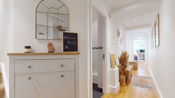 Un couloir avec une commode blanche, un miroir, une pancarte de bienvenue et une porte donnant sur une autre pièce.