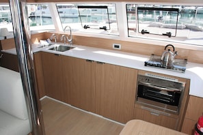 A compact galley kitchen with a sink, stove, and oven, wooden cabinetry, and large windows overlooking a marina. - Dubrovnik Delight: A Sun-Kissed Sailing Experience (Komolac)