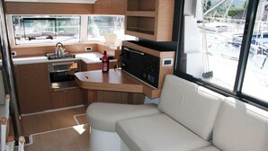 A compact kitchen with a stove, oven, and sink, a cushioned seating area, and a window with a view of a marina. - Dubrovnik Delight: A Sun-Kissed Sailing Experience (Komolac)