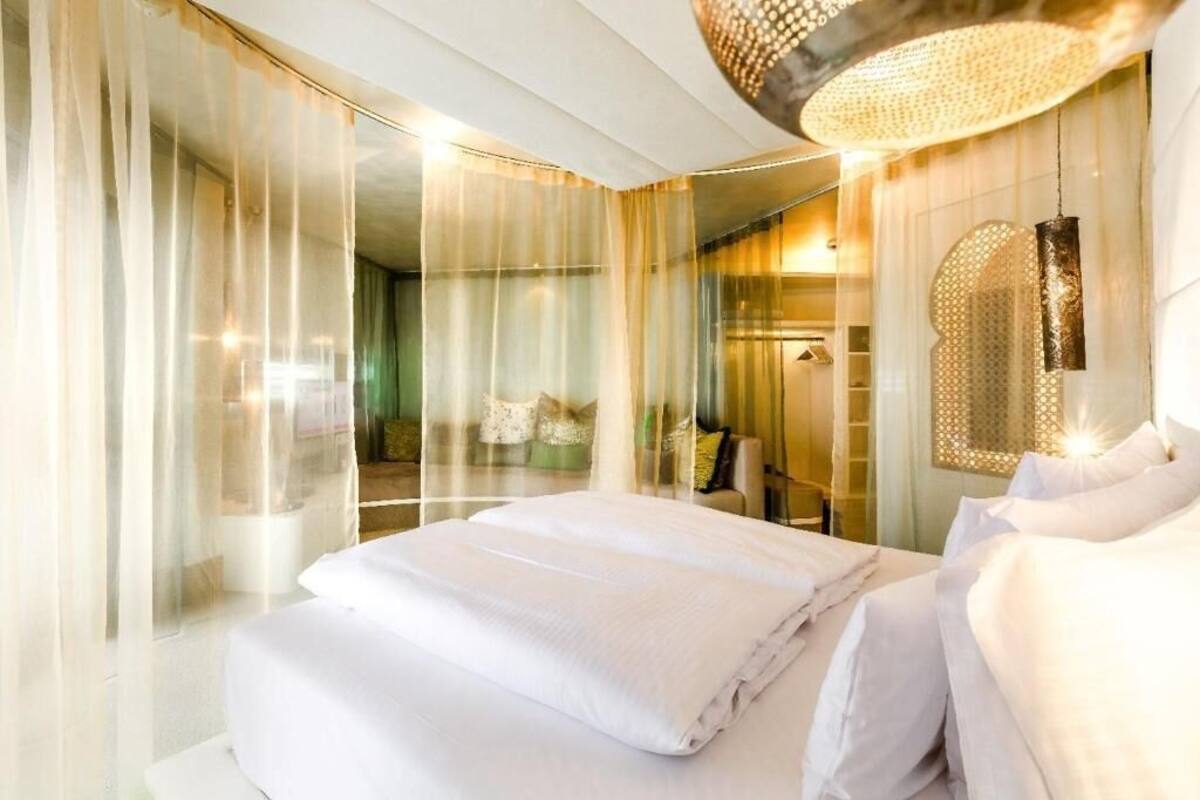 A hotel room with a large bed, a chandelier, and a view through sheer curtains.