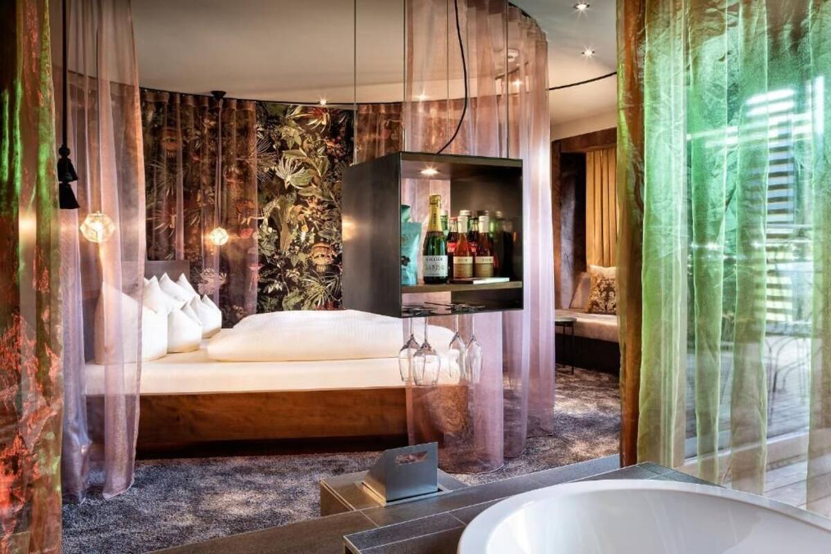 A modern bedroom with a bed, a minibar, a laptop, and a large window with sheer curtains.