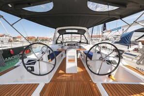 Een dek van een boot met een stuurwiel, een centrale tafel en uitzicht op andere boten en gebouwen.