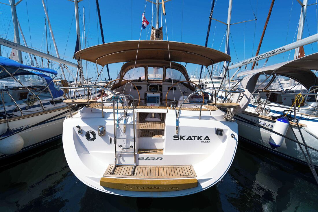 En vit yacht vid namn ”SKATKA” låg förtöjd i en småbåtshamn tillsammans med andra båtar. Den hade en hytt med en kontrollpanel och ett blått solskydd.