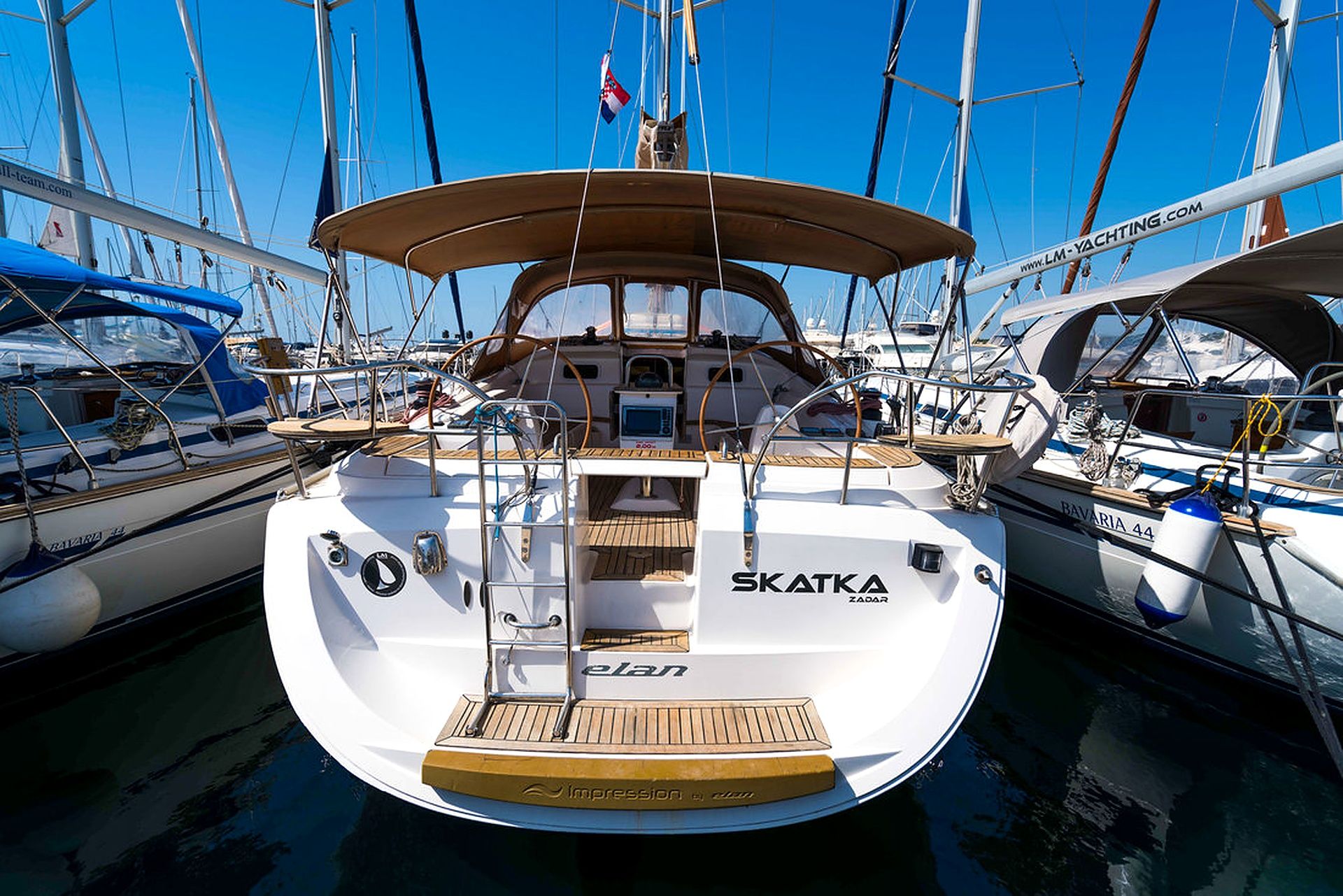 Un iat blanc anomenat 'SKATKA ZADAR' amarrat al port esportiu amb altres embarcacions, amb coberta de fusta i estructura de cabina.