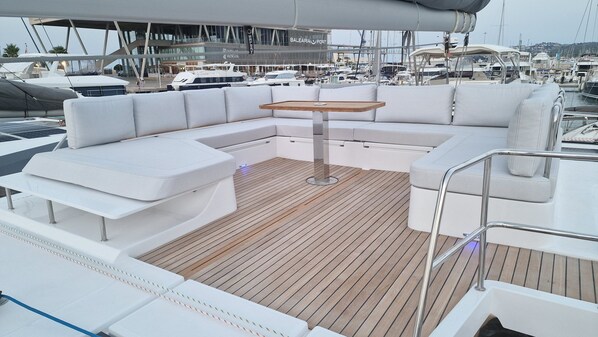 Un pont de bateau avec un espace salon doté de sièges blancs, une table en bois et une vue sur une marina avec des bateaux et un bâtiment portant l’inscription « BALEARIA PORT ».